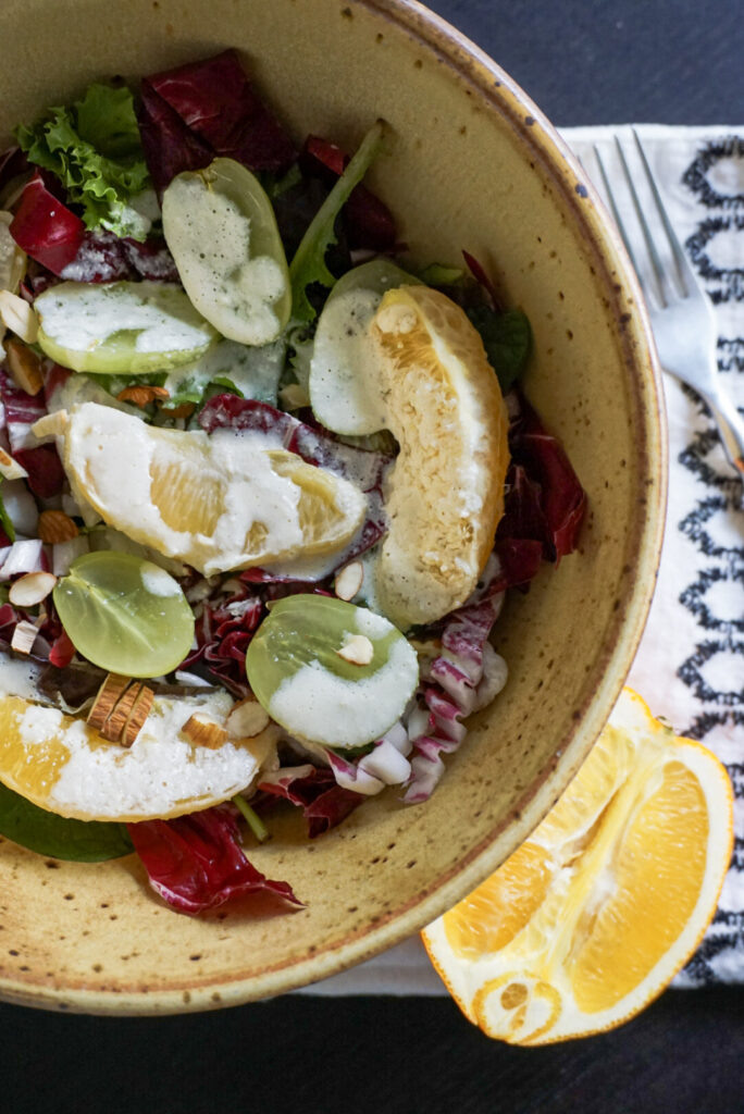 Orange wedges, green grapes, thinly sliced radicchio and a salad mix fill a large, yellow bowl that sits on top of a white and black place mate on top of a black, wooden table. The bowl sits next to a sliced orange.