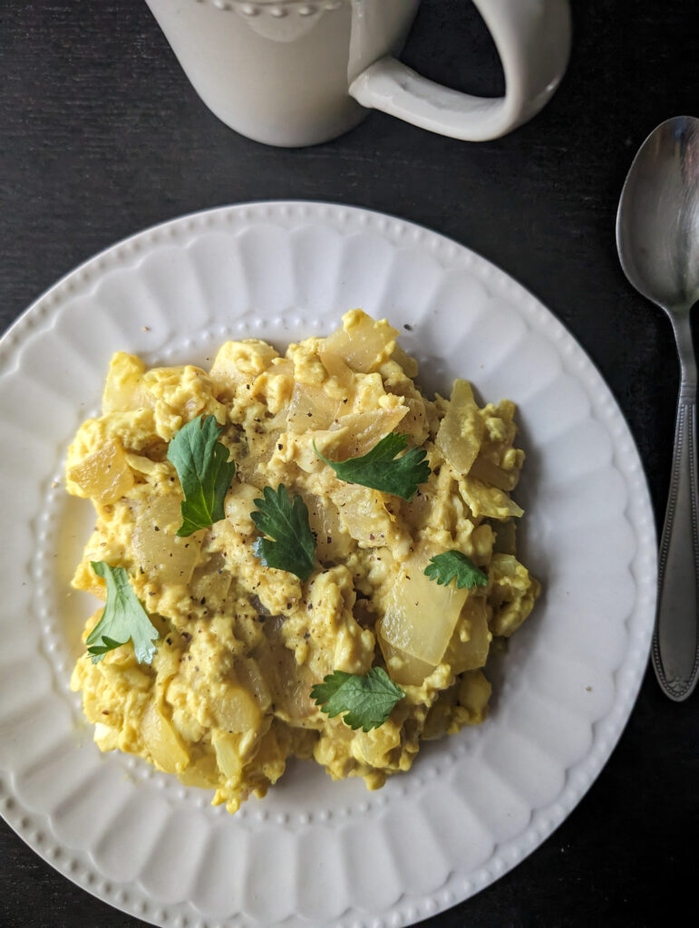 Yellow, vegan tofu scramble sits on a white plate on top of a black table. The scramble is topped with green parsley.