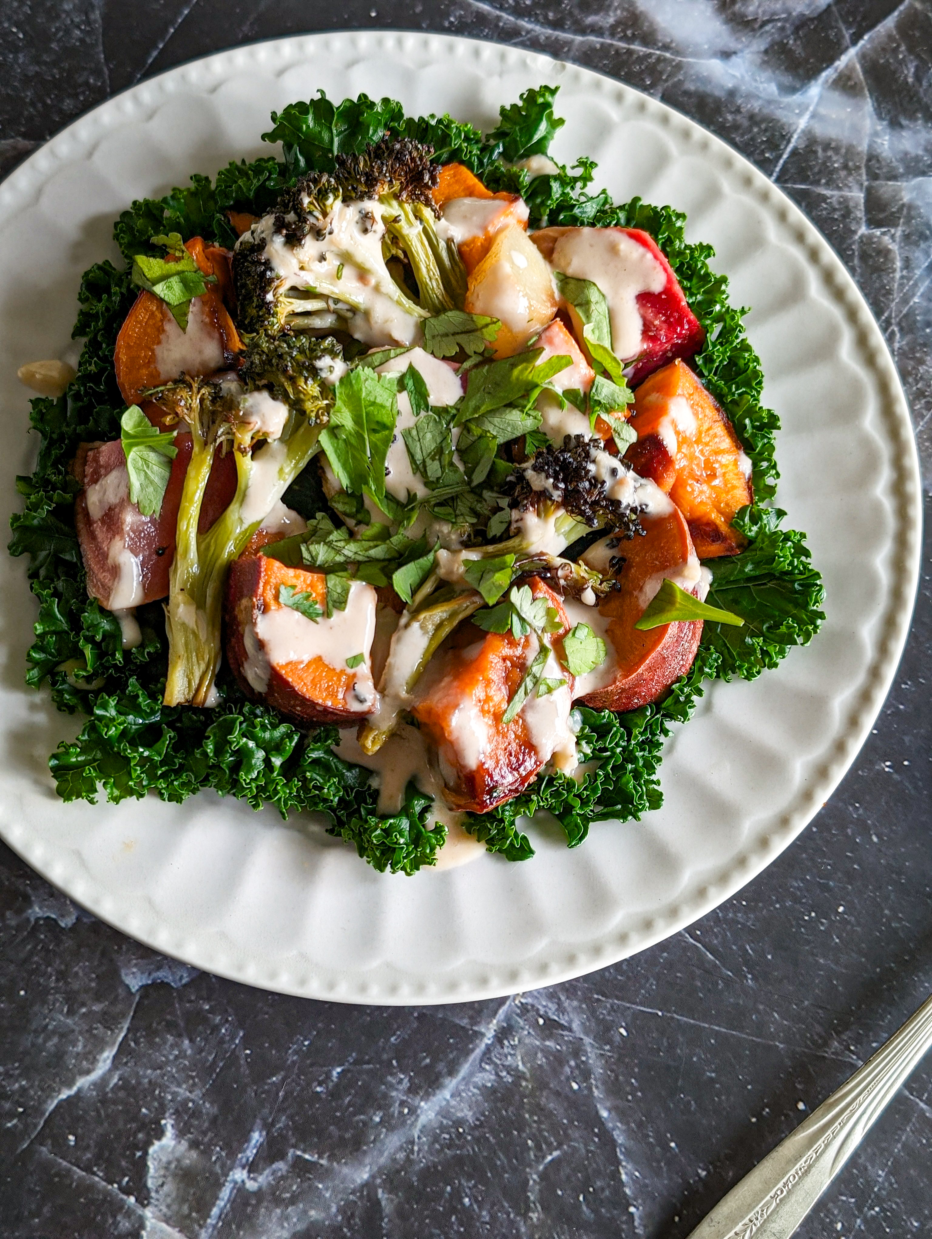 A variety of colorful, roasted vegetables including sweet potato and broccoli sit on a bed of steamed kale on top of a white plate, which sits on top of a gray and white marble counter top. The handle of a silver utensil sits near the bottom left corner of the plate.